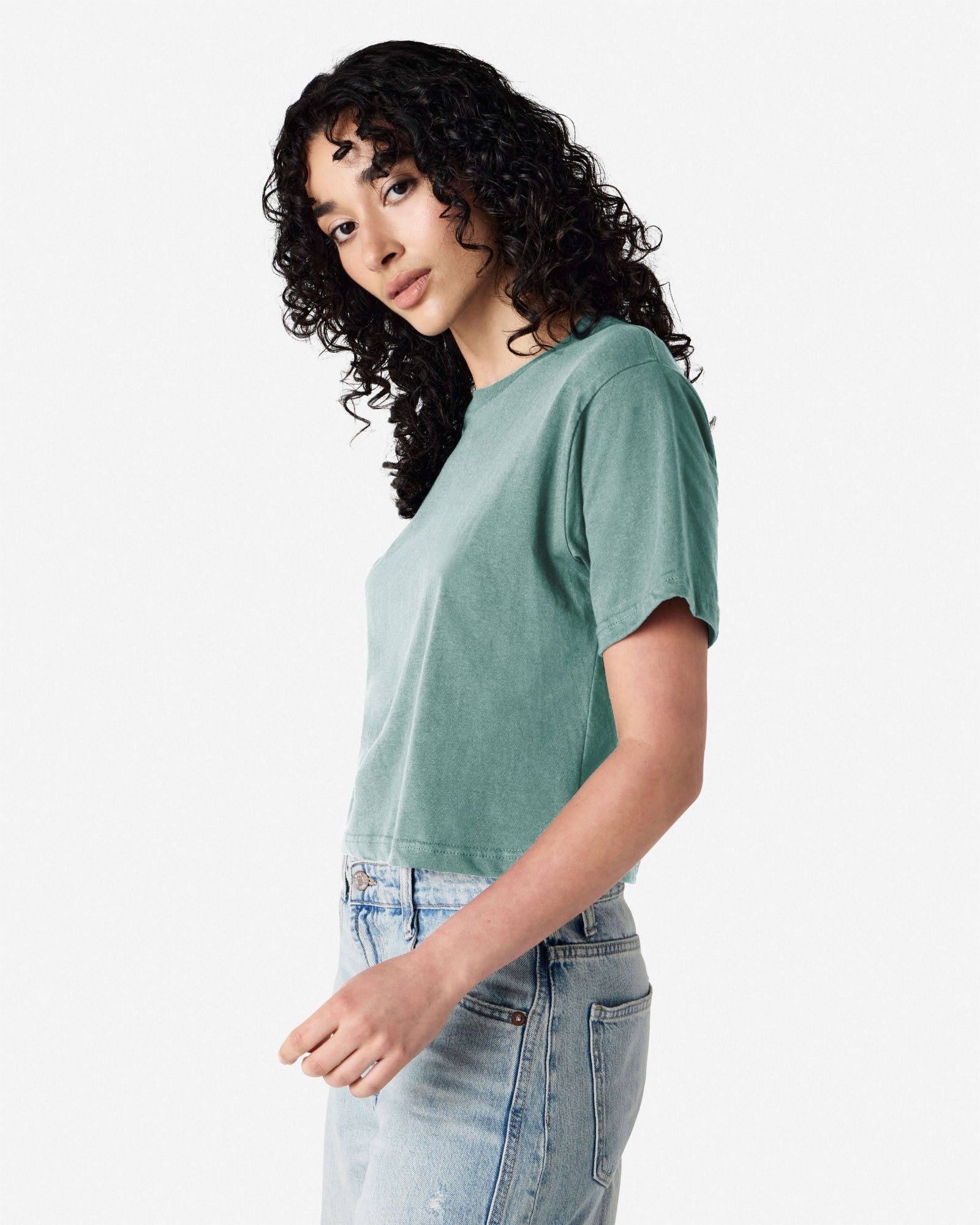 Female model wearing an American Apparel Fine Jersey Women's Boxy Tee in arctic, paired with blue jeans, standing against a plain background (side pose) -arctic