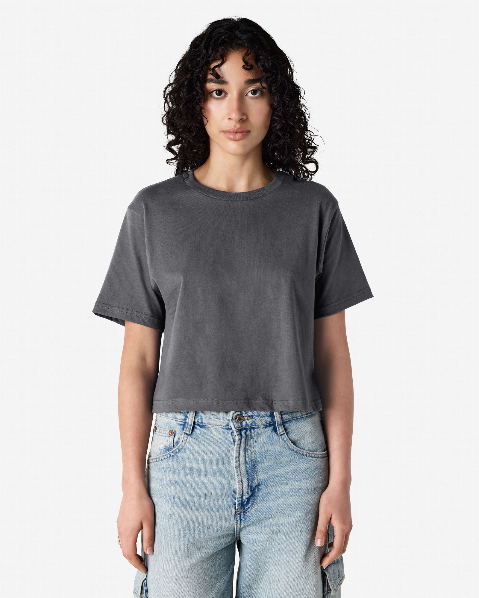 Female model wearing an American Apparel Fine Jersey Women's Boxy Tee in Asphalt, paired with blue jeans, standing against a plain background (front pose) -asphalt