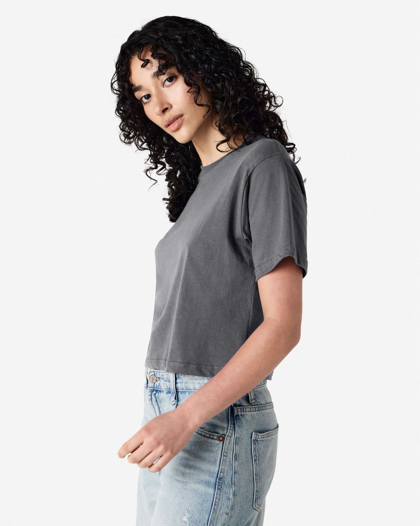 Female model wearing an American Apparel Fine Jersey Women's Boxy Tee in Asphalt, paired with blue jeans, standing against a plain background (side pose) -asphalt