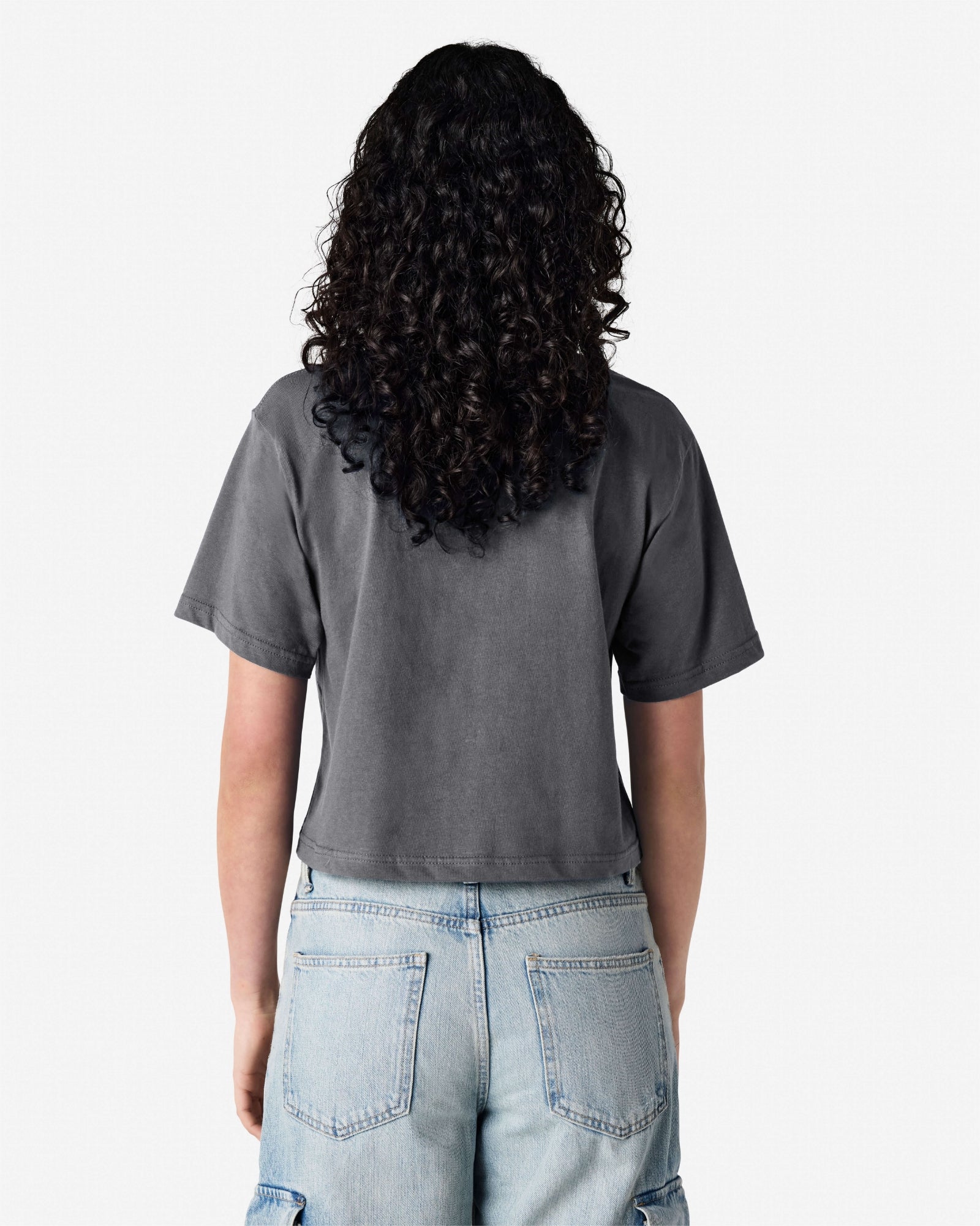 Female model wearing an American Apparel Fine Jersey Women's Boxy Tee in Asphalt, paired with blue jeans, standing against a plain background (back pose) -asphalt