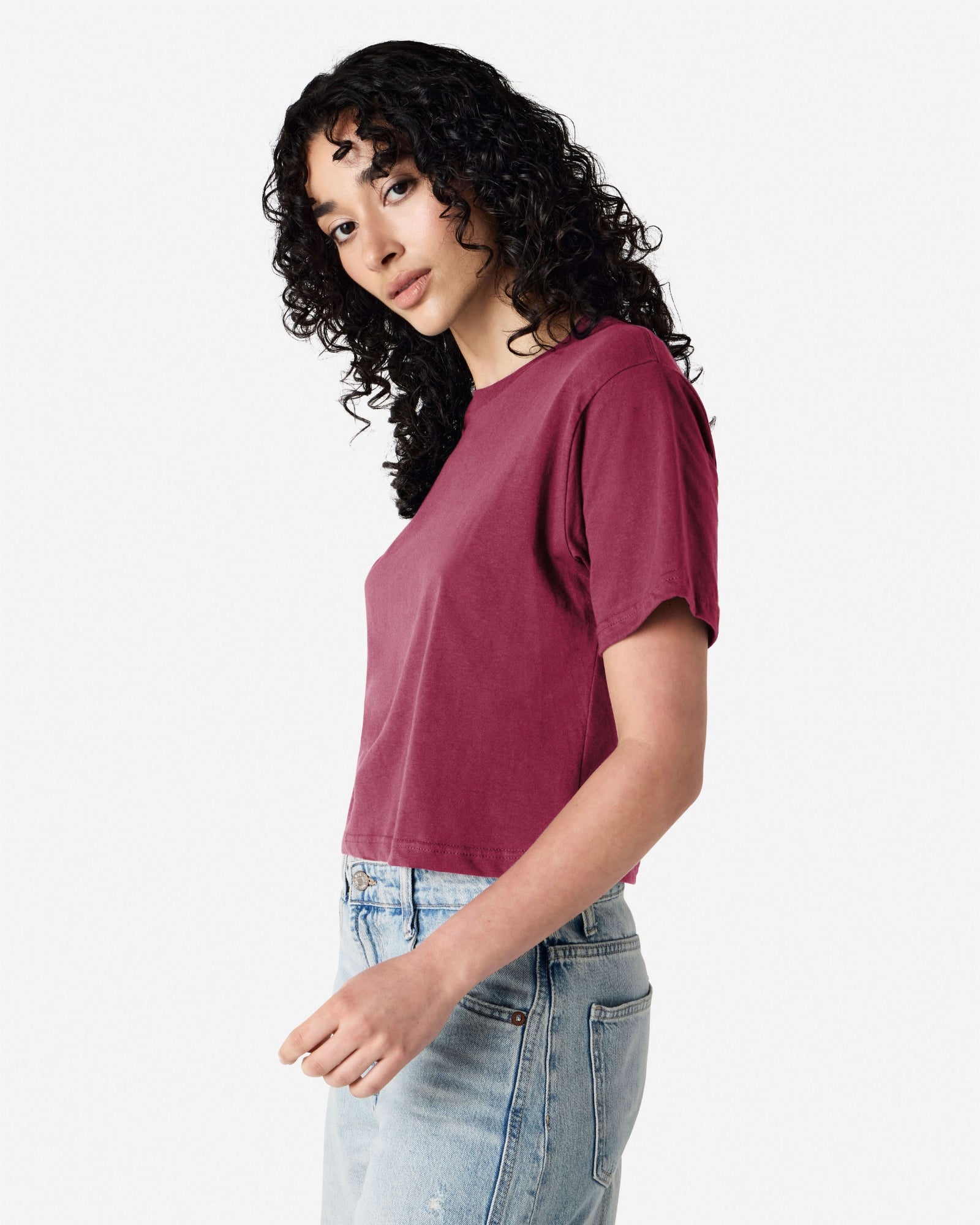 Female model wearing an American Apparel Fine Jersey Women's Boxy Tee in Burgundy, paired with blue jeans, standing against a plain background (side pose) -burgundy