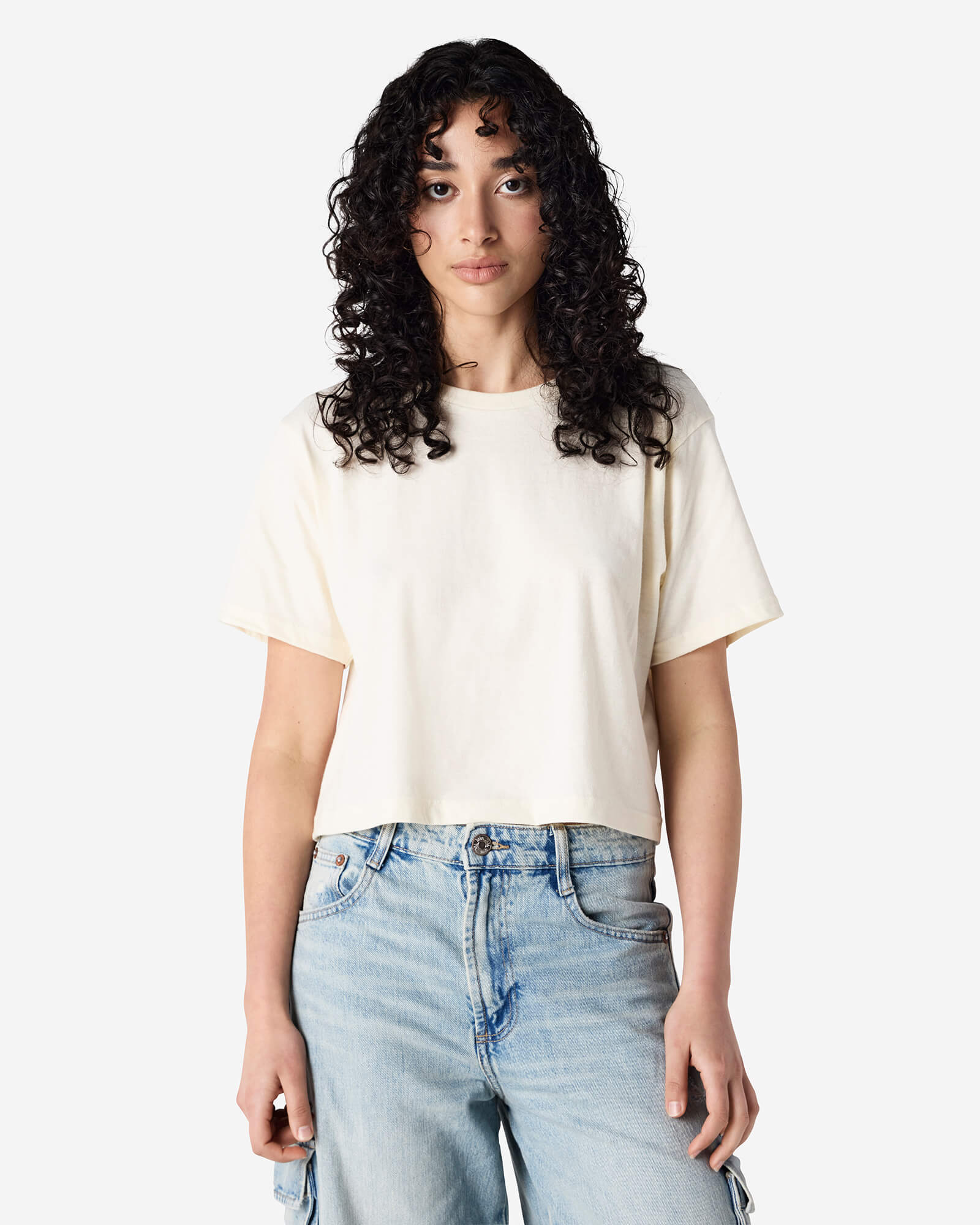 Female model wearing an American Apparel Fine Jersey Women's Boxy Tee in Cream, paired with blue jeans, standing against a plain background (front pose) -cream