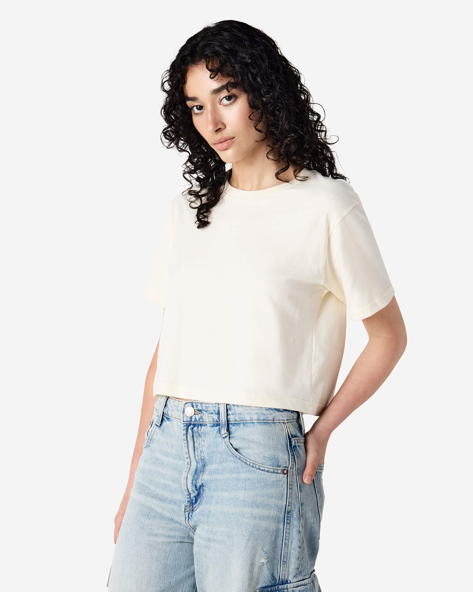 Female model wearing an American Apparel Fine Jersey Women's Boxy Tee in Cream, paired with blue jeans, standing against a plain background (front pose) -cream