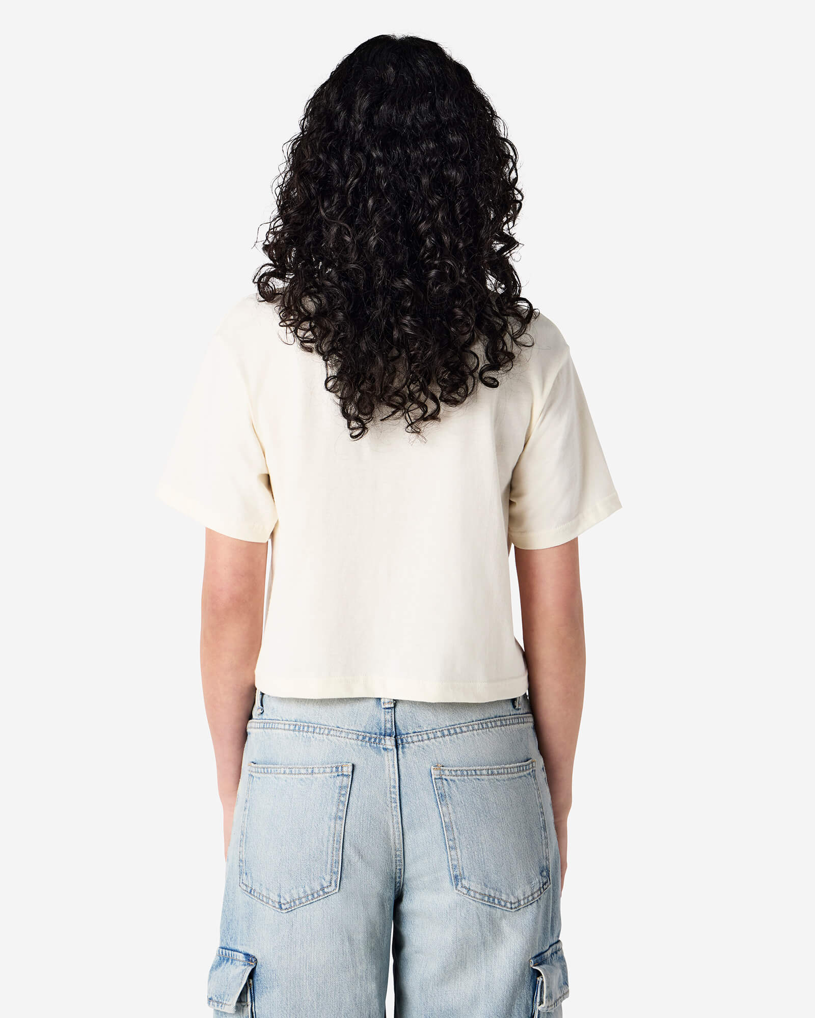 Female model wearing an American Apparel Fine Jersey Women's Boxy Tee in Cream, paired with blue jeans, standing against a plain background (back pose) -cream