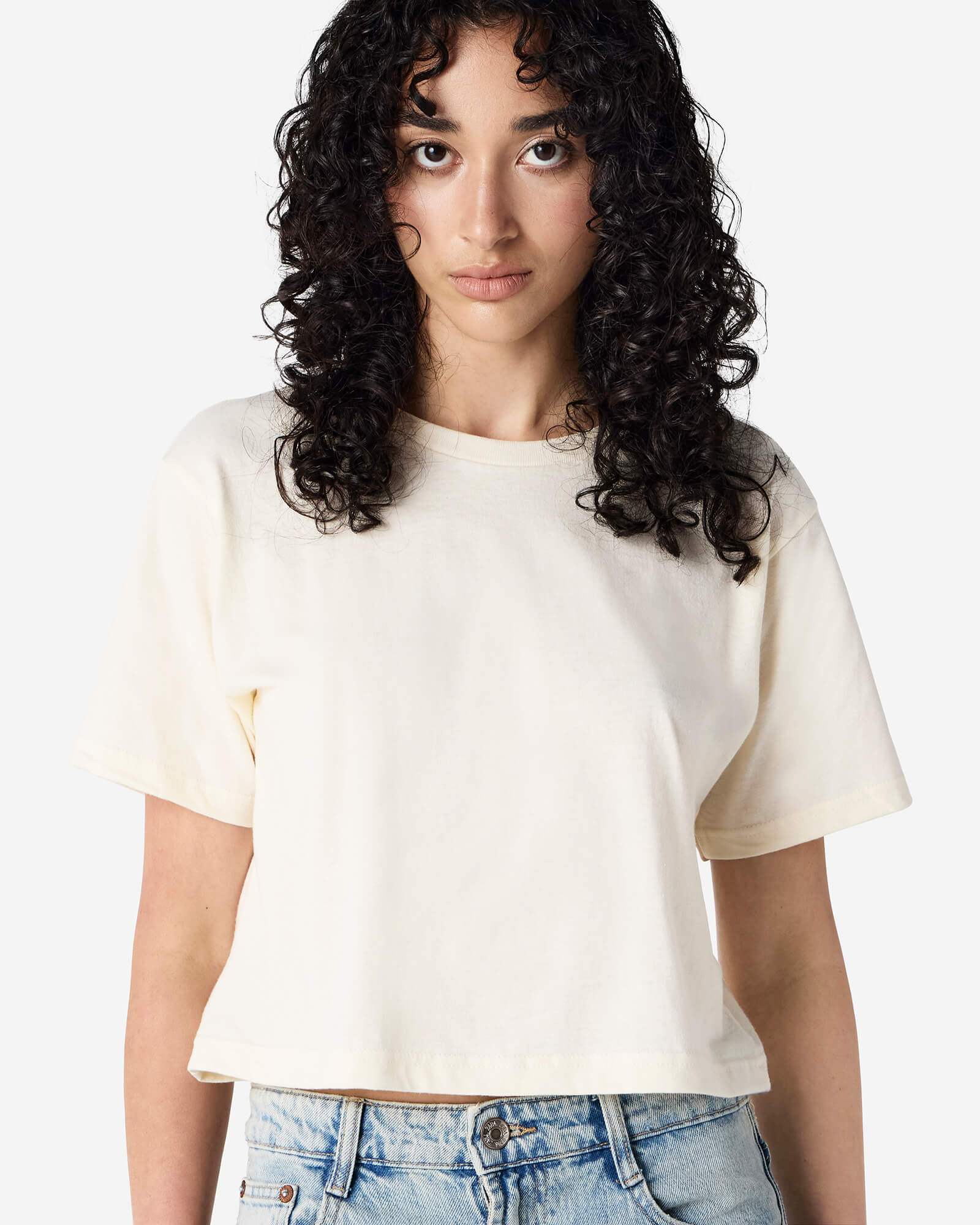 Female model wearing an American Apparel Fine Jersey Women's Boxy Tee in Cream, paired with blue jeans, standing against a plain background (front pose) -cream