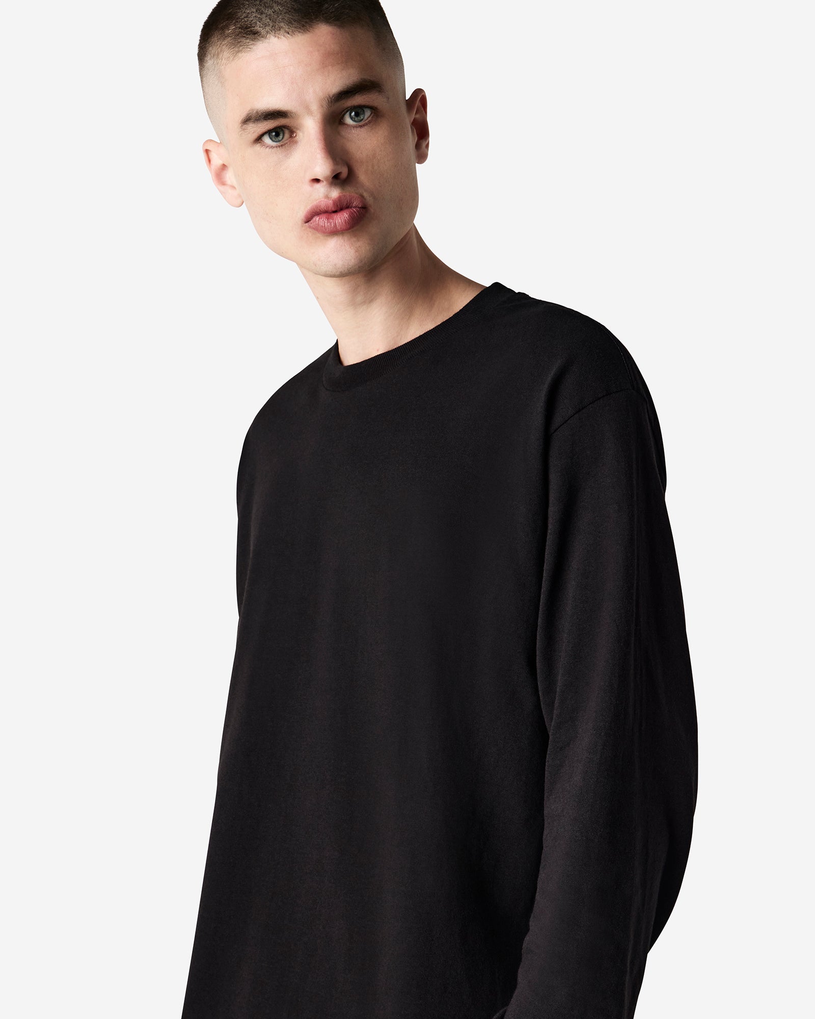 front view of a male Model wearing a black coloured Heavyweight Unisex Long Sleeve Crew Neck Tee, paired with blue jeans, standing against a plain background (front pose) -black