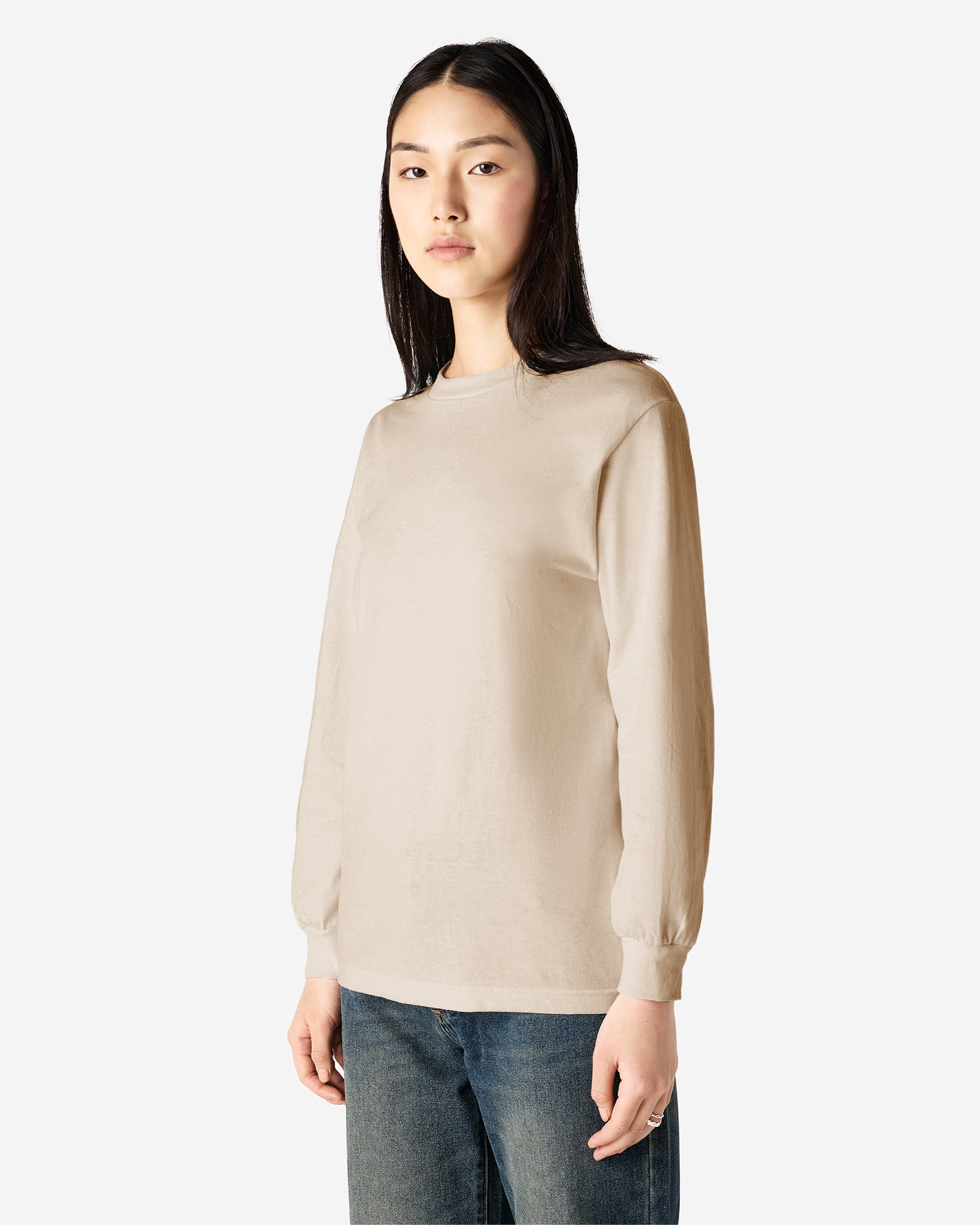 front view of a female Model wearing a Sand colored Heavyweight Unisex Long Sleeve Crew Neck Tee, paired with blue jeans, standing against a plain background (front pose) -sand