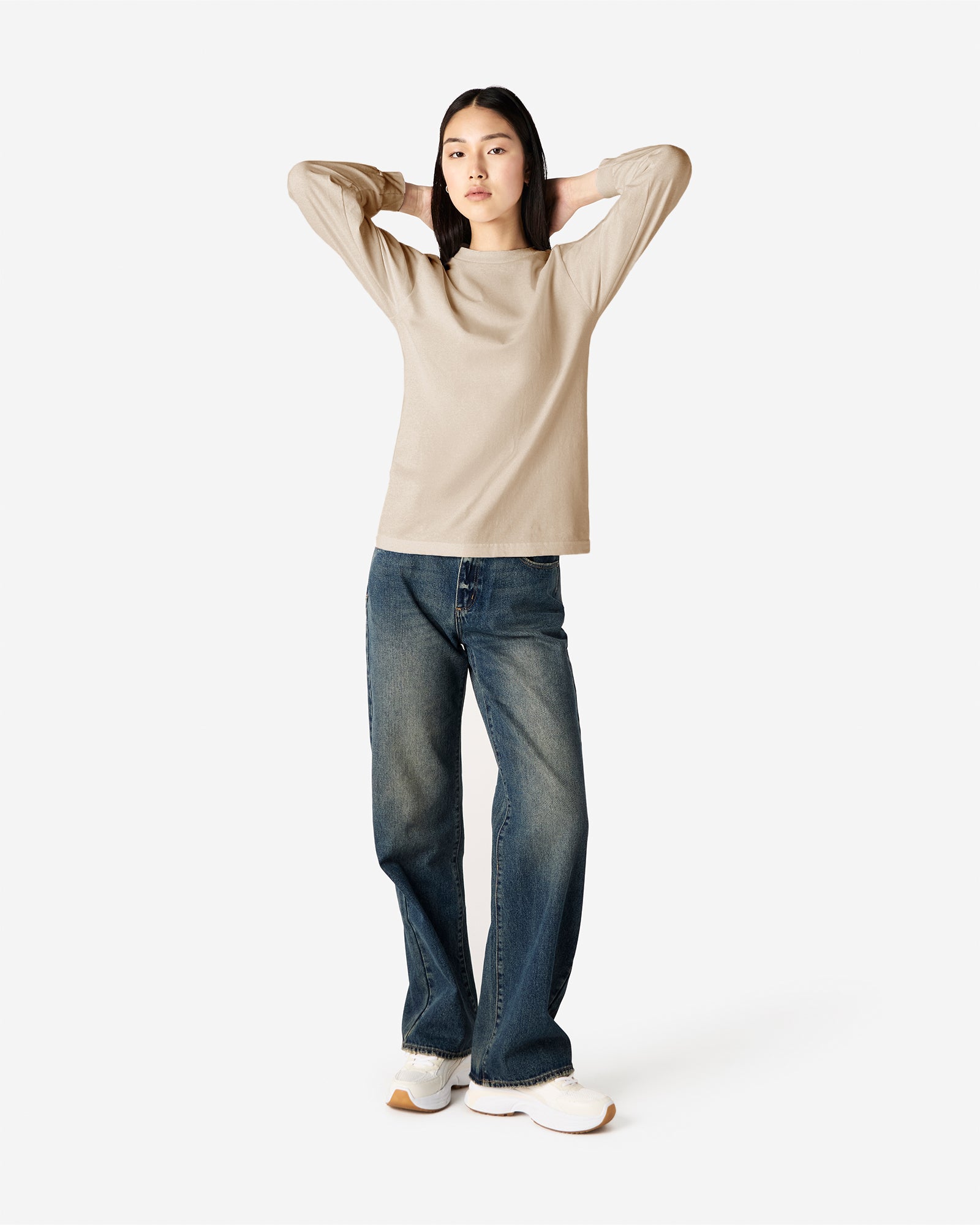 front view of a female Model wearing a Sand colored Heavyweight Unisex Long Sleeve Crew Neck Tee, paired with blue jeans, standing against a plain background (front pose) -sand